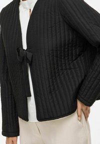 Svart vadderad jacka med rosettstängning, med texturerad finish, rundad krage och kortärmad design, kombinerad med en ljus topp och byxor.