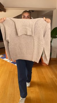 Beige kötött pulóver bordás nyakkal és ujjakkal, laza szabással és közepes hosszúságú testtel, egy személy mellett tartva.