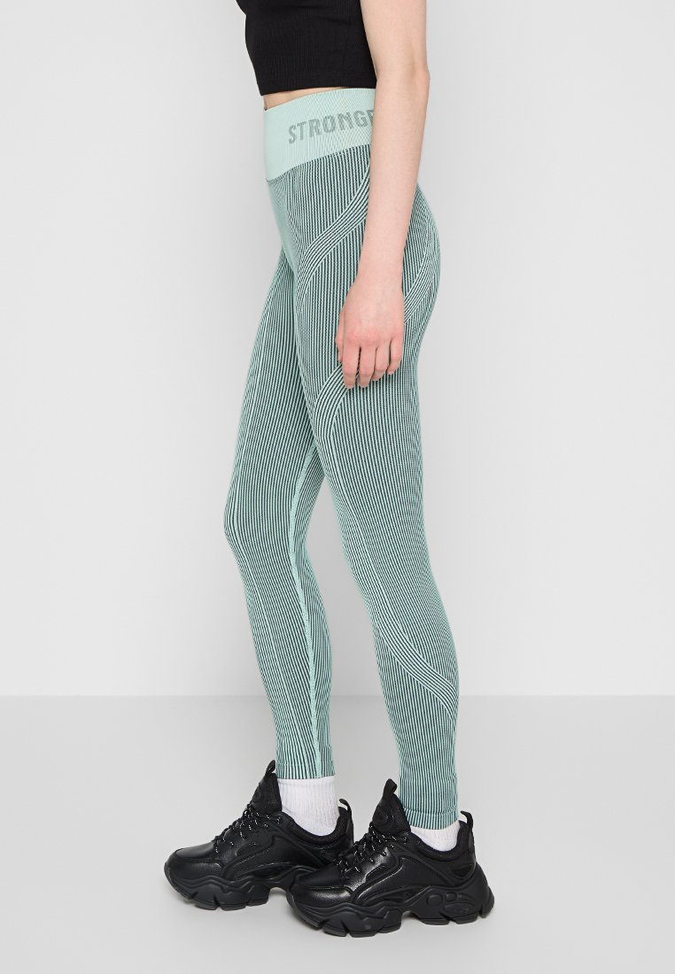 Zielone legginsy w paski z wysokim pasem z napisem "STRONGER". Materiał wydaje się elastyczny i gładki w dotyku. Noszone z czarnymi sneakersami.