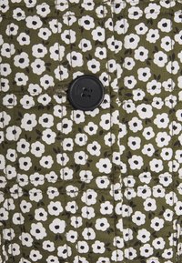 Bouton noir cousu sur un tissu matelassé vert olive avec de petites fleurs blanches aux centres noirs en motif répétitif.