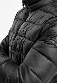 Giacca a vento nera con un design trapuntato, chiusura con zip, colletto alto e materiale resistente. Include una toppa con logo ricamato sul braccio.