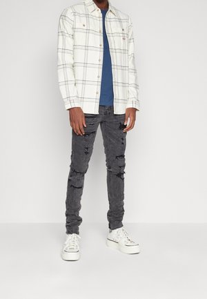 Chemise à carreaux blanche à manches longues sur un t-shirt bleu, associée à un jean noir déchiré et des baskets montantes blanches.