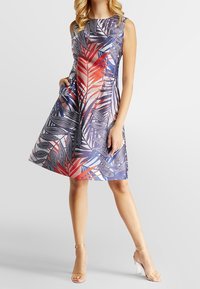 Robe à imprimé floral mettant en avant des couleurs bleue, rouge et blanche, sans manches, avec une jupe évasée et des poches latérales. Mannequin portant des talons transparents.