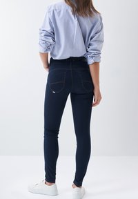 Granatowe wąskie dżinsy z tylnymi kieszeniami i pomarańczowymi przeszyciami, zestawione z jasnoniebieską koszulą na guziki i białymi sneakersami.