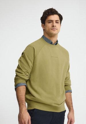 Uomo con felpa verde oliva sopra una camicia blu con colletto, in piedi con le braccia rilassate davanti a uno sfondo chiaro.