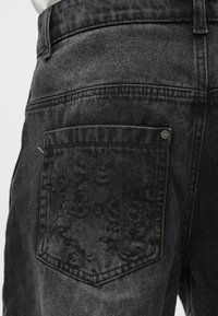 Temno sive jeans hlače iz jeansa z teksturiranim zadnjim žepom, okrašenim z vezenimi vzorci in kovinskim gumbom. Mehka, rahlo obrabljena površina.