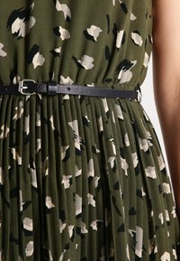 Vestido plisado verde con un patrón abstracto en crema y negro, ajustado en la cintura con un cinturón de cuero negro que cuenta con una hebilla plateada.