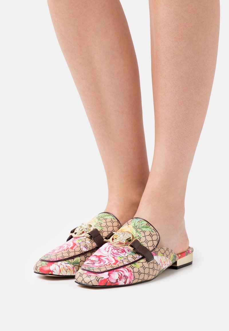 Pieds portant des mocassins à enfiler à motif floral avec des ornements dorés en forme de tête d’animal et des sangles marron, debout sur fond blanc.