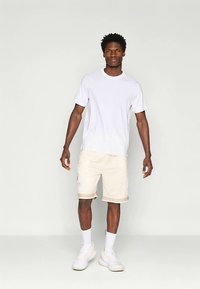 T-shirt de algodão branco combinado com calções creme com bolsos laterais e um emblema, usado com ténis e meias brancas.