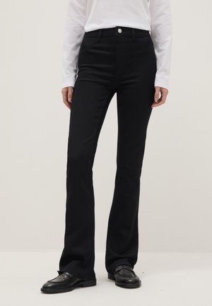 Personne portant un pantalon noir évasé taille haute, un haut blanc à manches longues, et des mocassins noirs, debout devant un fond clair uni.