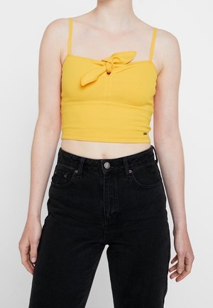 Kvinde iført en gul cropped tanktop med knude foran og sorte bukser med høj talje mod en ensfarvet baggrund.