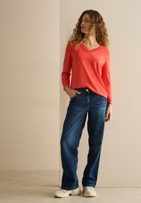 Korallenfarbener V-Ausschnitt-Pullover aus weichem Stoff, kombiniert mit weit geschnittenen, dunklen Jeans und weißen Sneakers, vor einem neutralen Hintergrund.