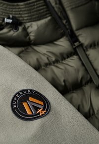 Zielona kurtka puchowa z gładkiego materiału o miękkiej fakturze, z okrągłą naszywką w czarne i pomarańczowe kolory z napisem "SUPERDRY."