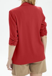 Punainen bleiseri, jossa on sileä textura, varustettuna kerätystä hihasta ja klassisesta kauluksesta, yhdistettynä khaki-shortsien kanssa.