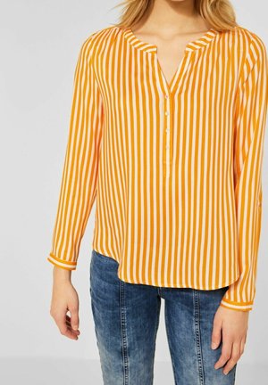 Vrouw draagt een blouse met lange mouwen, oranje-witte verticale strepen, een V-hals met knopen, gecombineerd met een blauwe spijkerbroek.