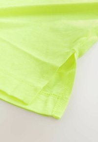 Neon gul bomulds-T-shirt, blød tekstur, korte ærmer og en lige kant, med synlige syningsdetaljer langs kanterne.