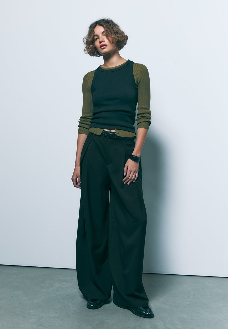 Črne široke hlače v kombinaciji s prilegajočo črno in olivno zeleno rebrasto majico. Obleka ima pas z naramnicami in gladko teksturo.