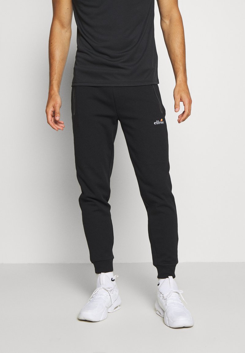 Black sweatpants made from soft fabric, featuring a tapered design, elastic cuffs, and a small logo on the left thigh. Paired with white sneakers.