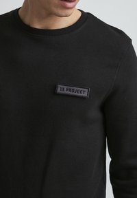 Black sweatshirt made of cotton with a round neckline. Features a square, woven label with embossed text "IL PROJECT" on the left chest area.