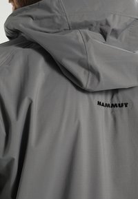 Personne portant une veste à capuche grise avec le logo "MAMMUT" brodé dans le haut du dos, vue en gros plan de la texture du tissu et des coutures.