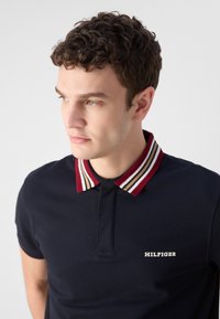 Ναυτικό πόλο μπλουζάκι με ριγέ γιακά σε κόκκινες, λευκές και μπεζ αποχρώσεις. Λογότυπο "HILFIGER" κεντημένο στο αριστερό στήθος. Βραχίονες κοντοί.