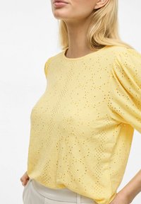 Haut jaune à manches courtes avec un motif en œillets texturé. Encolure ronde, manches légèrement bouffantes et tissu léger.