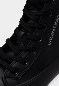 Μαύρο αθλητικό παπούτσι ψηλού τύπου με λείο δέρμα, στρογγυλά κορδόνια, μαύρες τρύπες κορδονιών και λεπτομέρεια λογότυπου στο πλάι. Υφή στη μόδα από καουτσούκ.