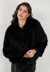Woman with wavy brown hair wearing hoop earrings, glossy lipstick, a black faux fur jacket, and black leather pants against a plain background.