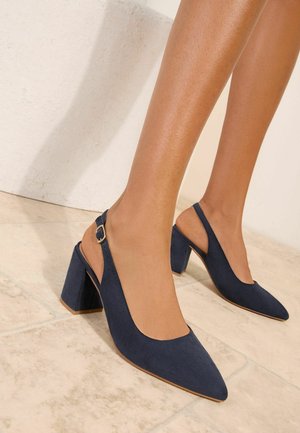 Jalad, mis kannavad tumelõhnavat sinist seemisnahast rihmikuga kontsakingi ninaosaga ja plokktaldadega, seisavad beeži plaatidega põrandal valge seina lähedal.