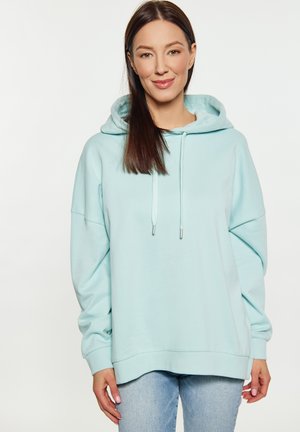 Felpa oversize azzurra chiara realizzata in un materiale morbido, con cappuccio regolabile e polsini a costine, abbinata a jeans chiari.