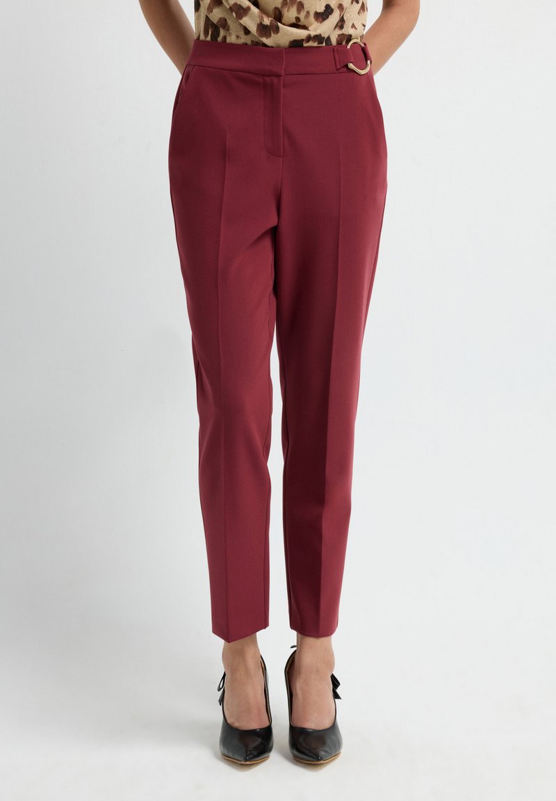Woman wearing high-waisted burgundy tapered pants with a belt detail, paired with black pointed-toe heels and a leopard print top.