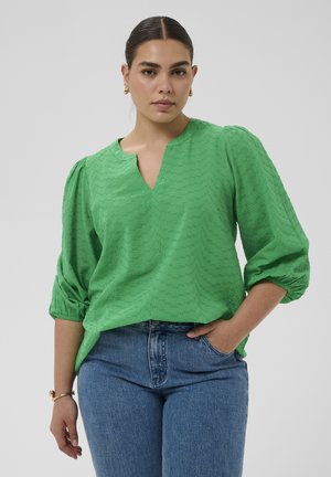 Kobieta ubrana w zieloną bluzkę o fakturze z rękawami bufkami do połowy przedramienia i niebieskie dżinsy, stojąca z jedną ręką w kieszeni na jednolitym tle.