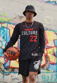 Fekete, hosszú ujjú mez piros "HEAT CULTURE 22" felirattal, fekete rövidnadrág Miami Heat logóval, kosárlabdát tartva, színes falfestmény előtt.