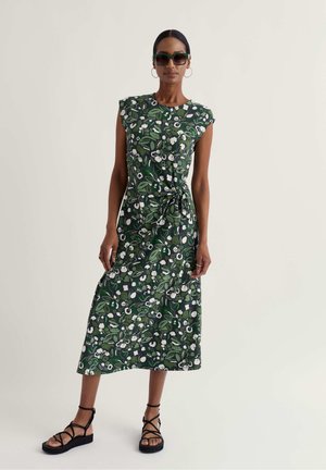 Femeie purtând o rochie midi verde cu imprimeu floral și talie cu șnur, sandale negre cu barete, cercei cerc și ochelari de soare întunecați, stând în picioare pe un fundal simplu.