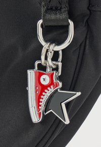 Γοητευτικό κόκκινο και λευκό αθλητικό παπούτσι προσαρτημένο σε ασημένιο μπρελόκ με μαύρο αστεράκι, πάνω σε υφή μαύρης επιφάνειας.