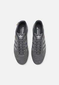 Szürke velúr sportcipők szürke és fekete csíkokkal, kerek orral, fűzős kialakítással és texturált talppal. Adidas logó a nyelvén.