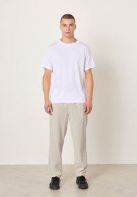 Jaunas vyras stovintis priešais šviesią paprastą foną, dėvintis baltą marškinėlius, smėlio spalvos kelnes ir juodus sportinius batelius, žvelgiantis tiesiai su neutraliu išraiška.