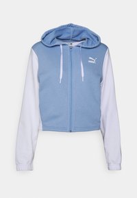 Haut de survêtement zippé coupé en bleu et blanc. Le matériau est doux, avec un corps bleu uni, des manches blanches et un logo sur la poitrine. Comprend une capuche.