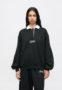 Γυναίκα με μακριά σκούρα μαλλιά φορώντας μαύρη φανέλα Jordan με λευκό γιακά και ταιριαστά μαύρα παντελόνια Jordan, μπροστά σε απλό λευκό φόντο.