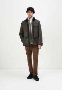 Giovane in giacca verde scuro sopra un maglione grigio a maglia, pantaloni marroni e scarpe nere, in piedi contro uno sfondo bianco semplice.