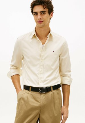 Ung mand iført beige skjorte med opkrogede ærmer, brune bukser og sort bælte, står med den ene hånd i lommen.