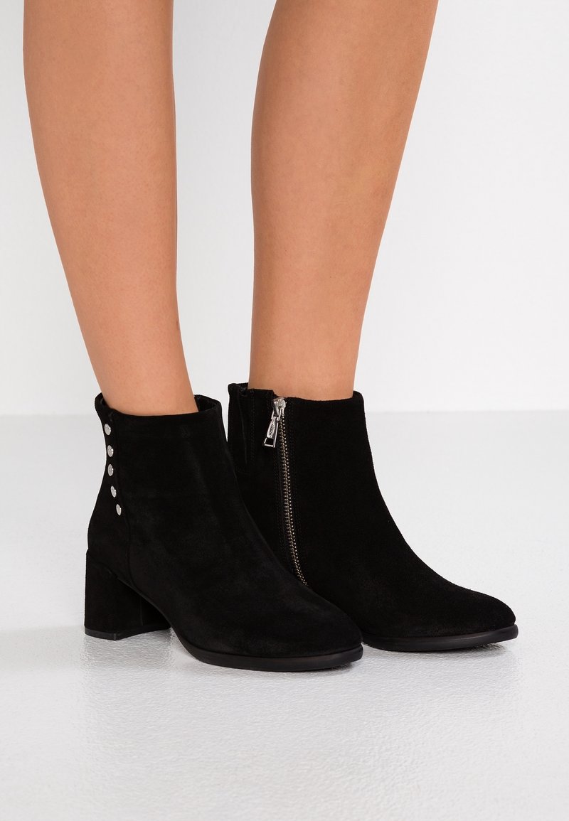 Bottines en daim noir à talons carrés, fermeture éclair latérale sur la botte droite, et boutons argentés décoratifs sur la botte gauche portées sur des jambes nues.