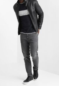 Veste en cuir noire avec épaules rembourrées, pull marine avec une bande blanche, jean gris, et baskets noires avec semelles blanches.