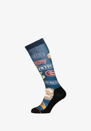 Blauwe kniekous met zwarte hiel en teen, met kleurrijke vintage-stijl patches en tekstzinnen zoals "PETST" en "Mountain Pioneers."