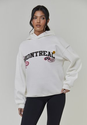 Biała bluza z kapturem z dużym napisem "MONTREAL", kolorowymi naszywkami i troczkiem przy kapturze. Długie rękawy z ściągaczami i luźny krój.