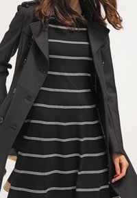 Zwarte trenchcoat met knopen en een kraag, laag over een zwart-wit gestreepte jurk met korte mouwen en een uitlopende rok.