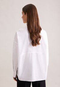 Camicia bianca oversize con orlo arrotondato, maniche lunghe e polsini con bottoni; tessuto liscio; cuciture dettagliate sul retro.