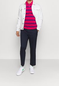 Bílá denimová bunda, růžové pruhované polo triko, tmavě modré kalhoty a bílé tenisky se zelenými akcenty. Neformální outfit se vrstvenými texturami.