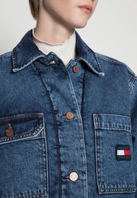 Denim jacket in dark blue with copper buttons, two front pockets, and a stitched logo patch featuring red, white, and green horizontal stripes.