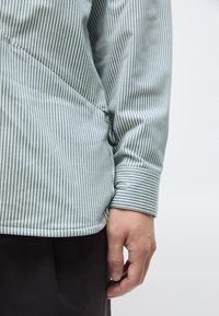 Striped skjorte i lyseblå og hvit bomull. Har en asymmetrisk kutt, glidelåsdetalj og knappet mansjett. Hånden hviler langs kanten.